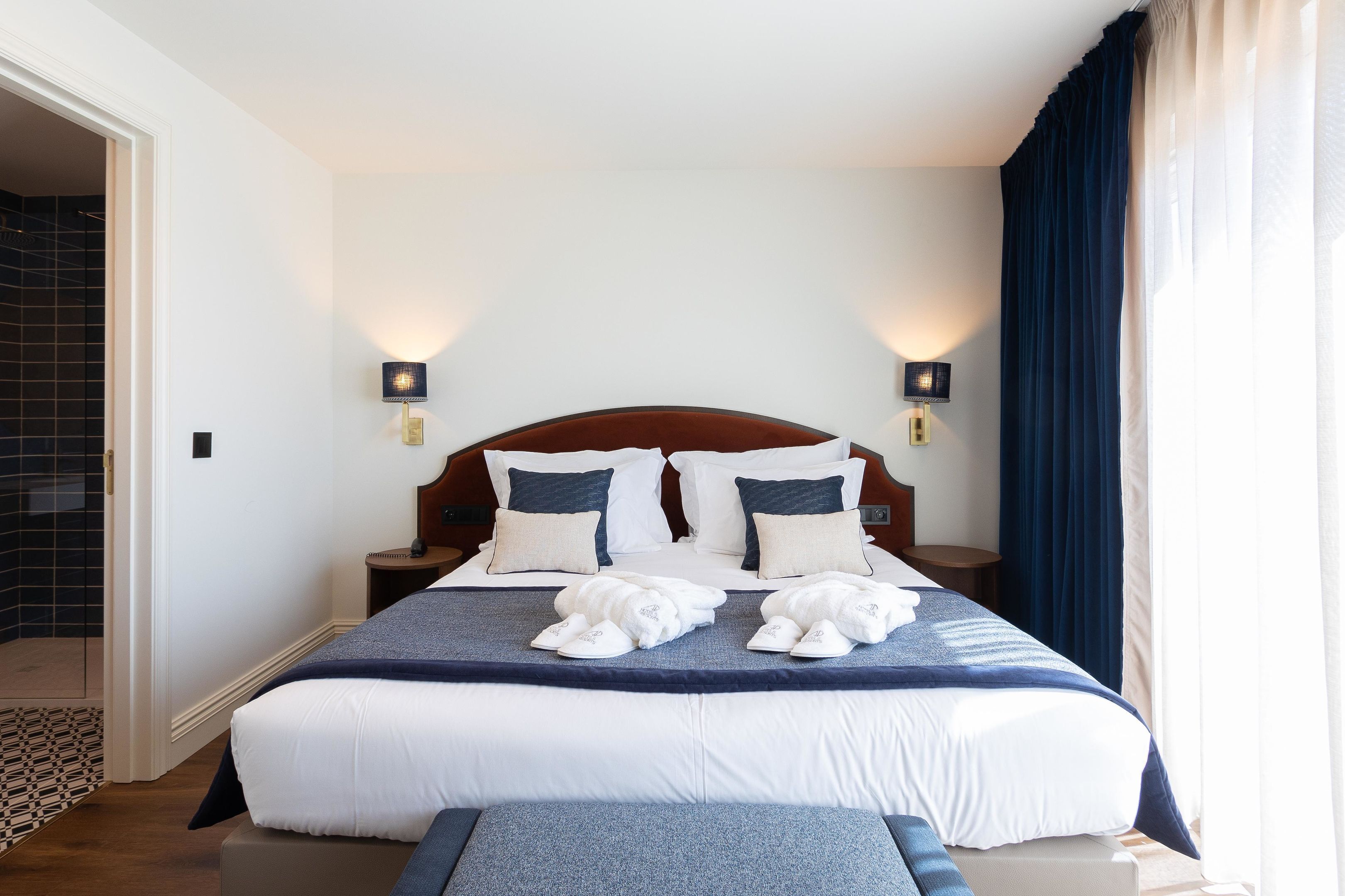 A neatly made bed with white and navy bedding, two bedside lamps, and a pair of slippers on the bed.