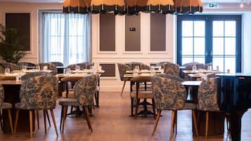 A dining area with multiple tables and chairs, a piano, and large windows with curtains.