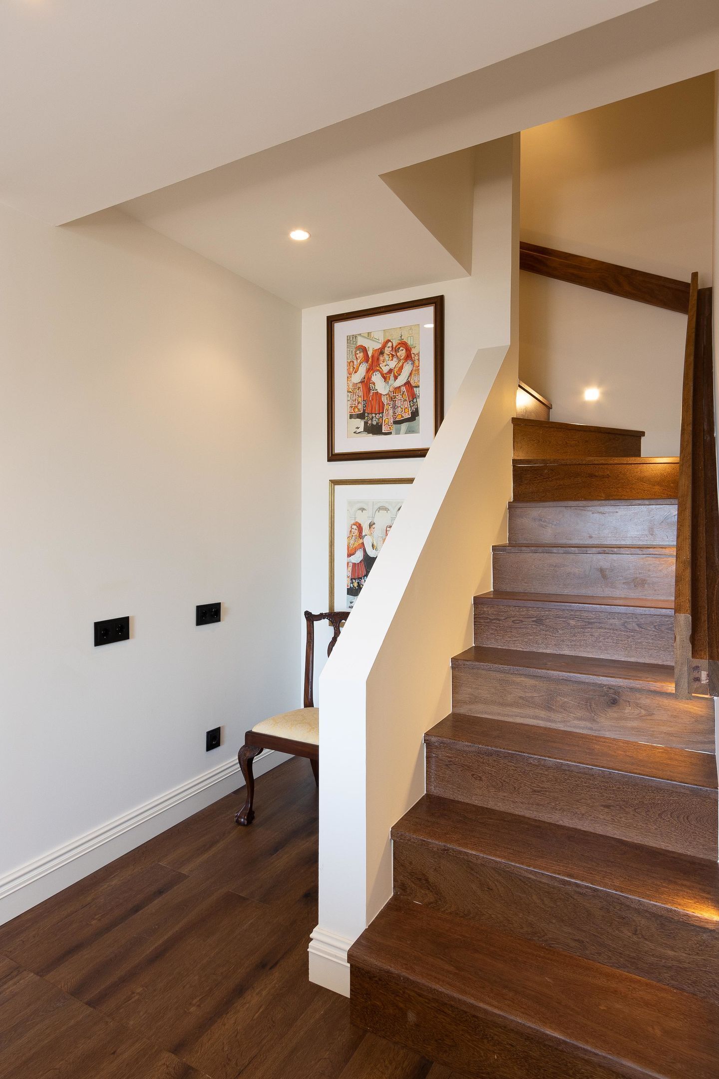 A wooden staircase with a chair at the bottom, white walls, and two framed pictures on the wall.