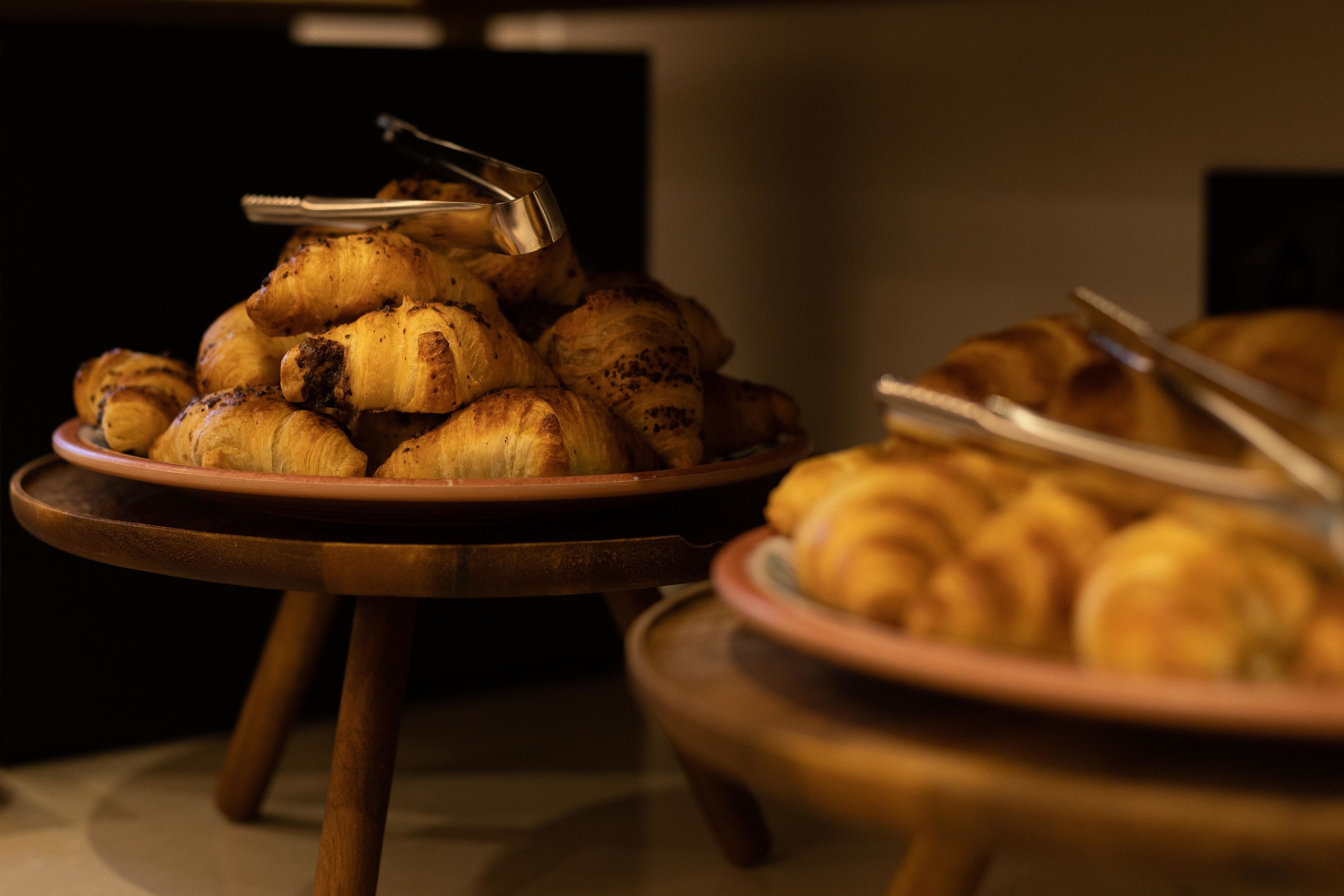 Una mesa de buffet con platos de pasteles, incluyendo cruasanes y otras preparaciones horneadas, con unas pinzas al lado.