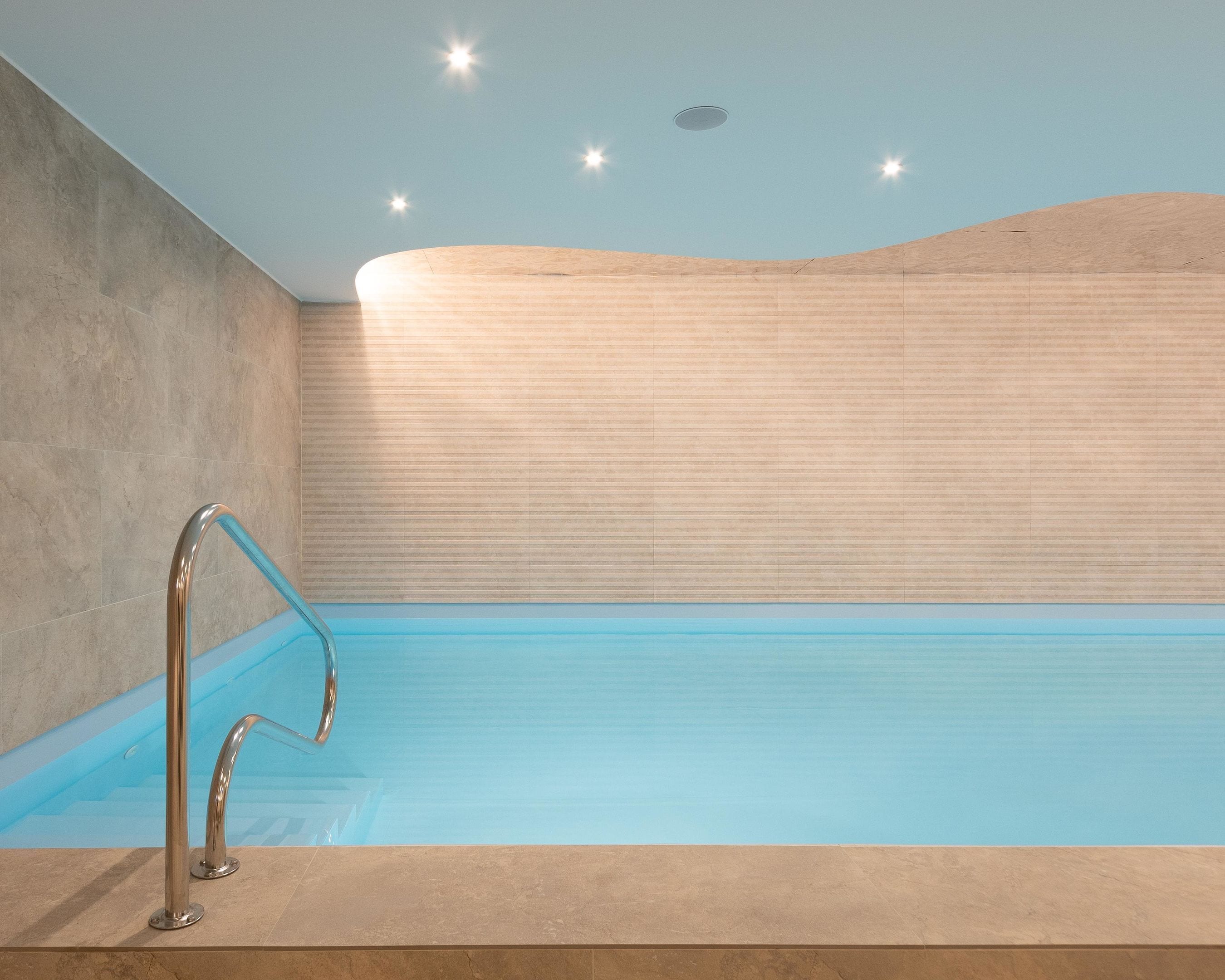 A modern indoor swimming pool with a clear blue water surface, a metal pool ladder, and a textured wall in the background.