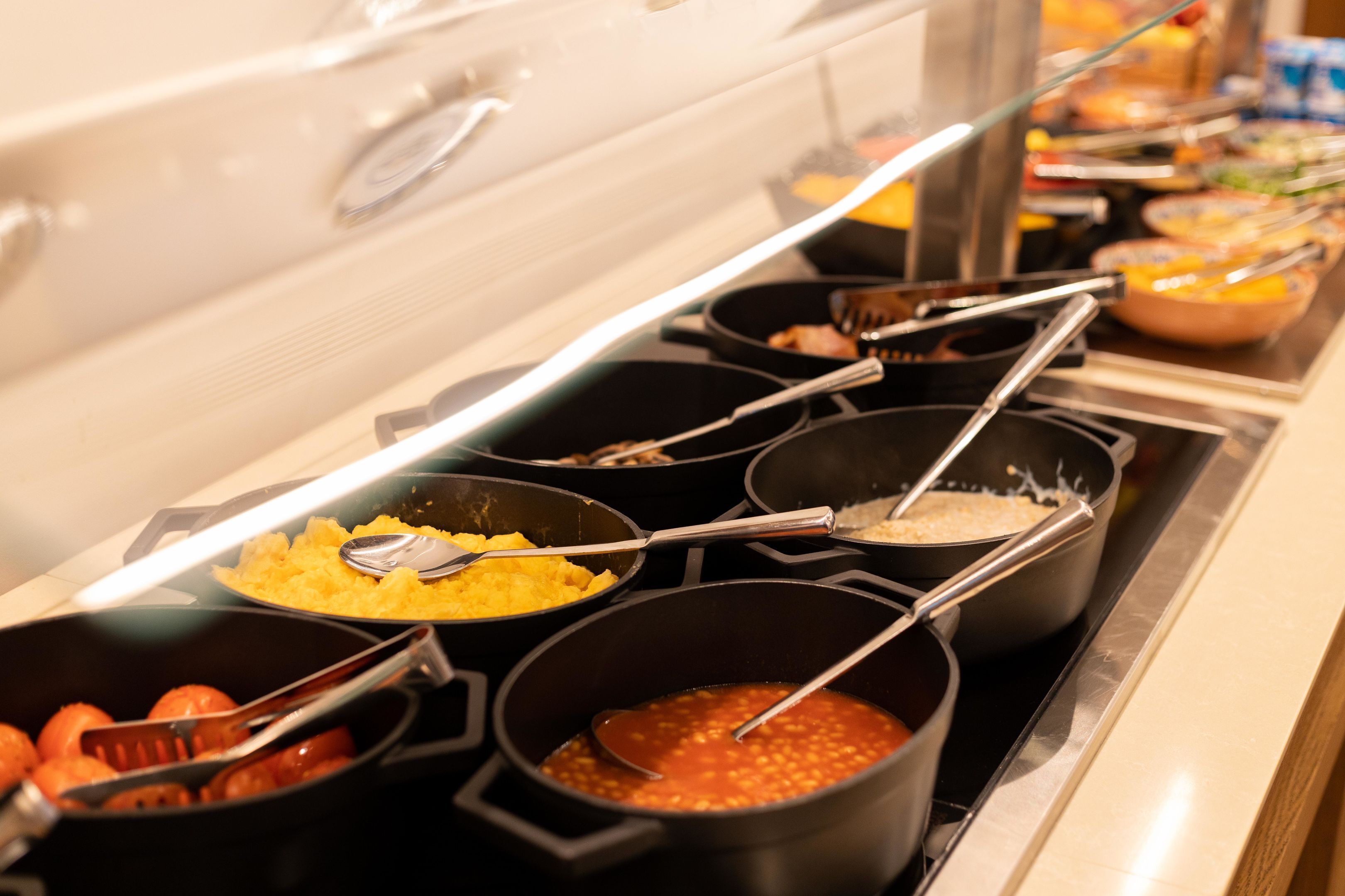 Una barra de desayuno con varios platos en ollas y recipientes, incluyendo aparentemente huevos revueltos, salsa roja y otros alimentos preparados.