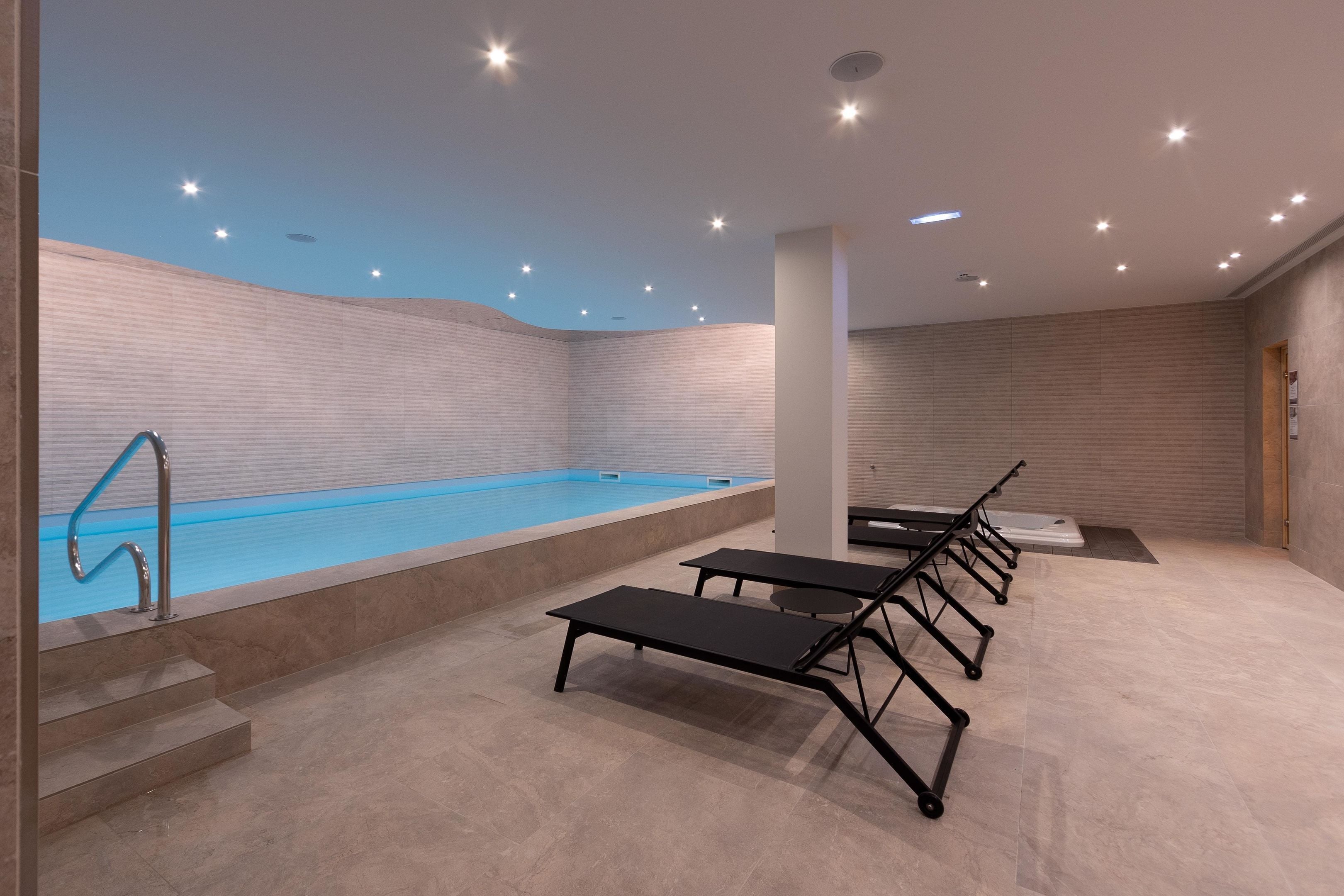 Un área de piscina interior moderna con una piscina azul cristalina, sillones de descanso negros y suelo de baldosas.