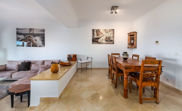 Een moderne woonkamer met een bank, een eettafel met stoelen en een klein eilandje met een vaas. Twee schilderijen hangen aan de muren en er staat een kast met een spiegel.