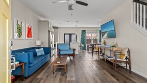 Amplia sala de estar con un sofá azul, una mesa de centro de madera y una mesa de comedor con sillas. Se observa un ventilador de techo, y hay cuadros enmarcados en la pared.
