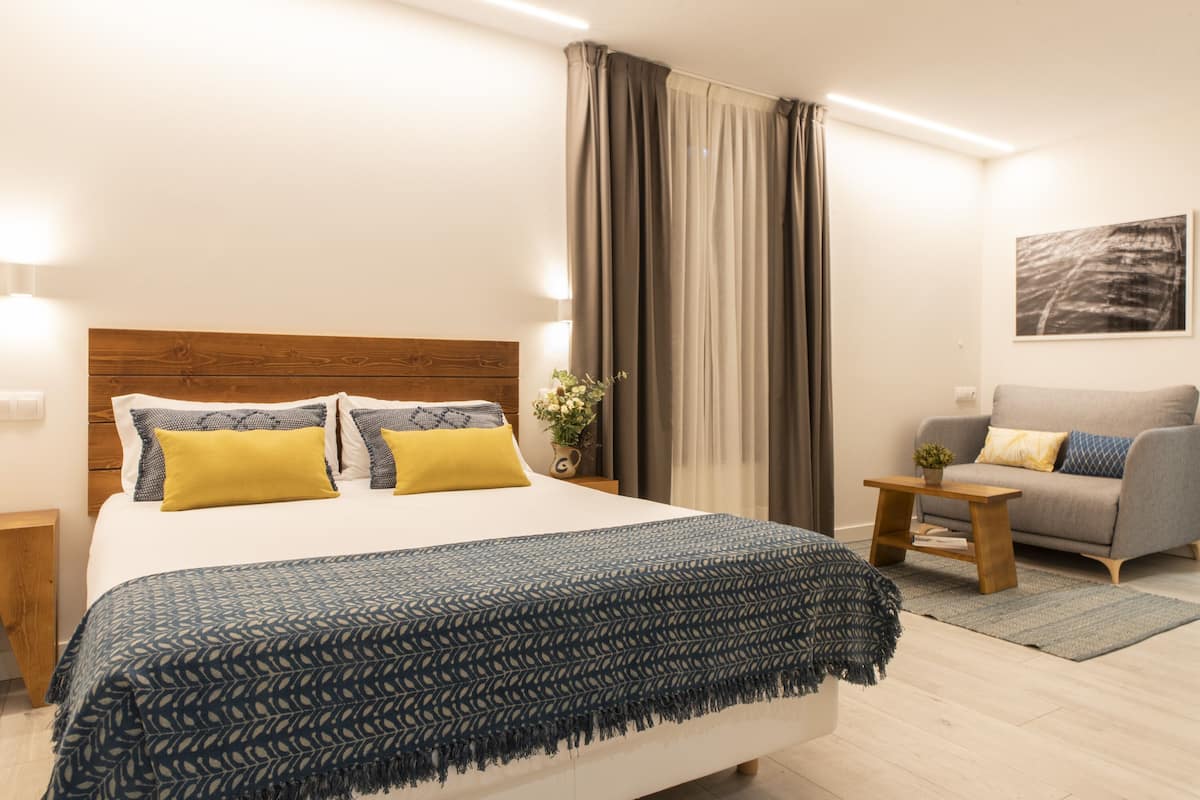 A modern bedroom with a large bed, a wooden headboard, a grey sofa, and a small table with a plant.
