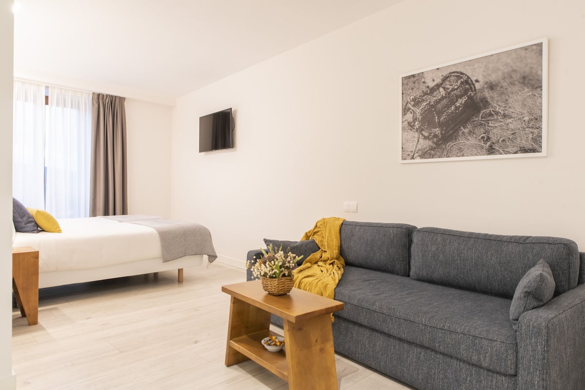A modern living room with a grey sofa, a wooden coffee table, a white bed with a grey and yellow pillow, and a black television on the wall.