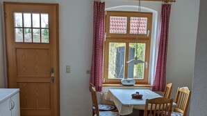 Una habitació amb sòl de fusta, una taula de dinar amb quatre cadires, una finestra amb cortines i una porta.