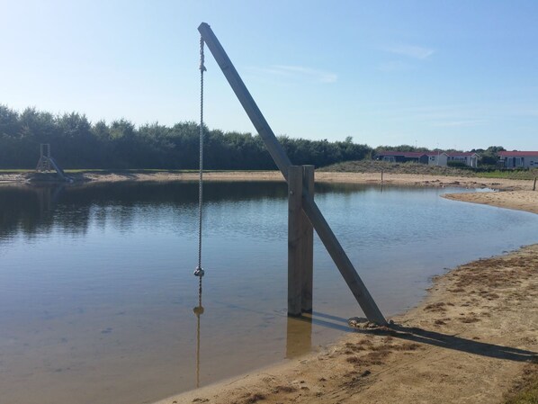 Una estructura de concreto junto a una masa de agua tranquila con orilla arenosa, rodeada de vegetación y algunos edificios al fondo.