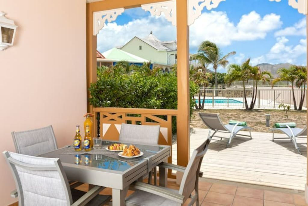 Comedor con mesa y sillas, zona de descanso junto a la piscina con tumbonas, y vista a una piscina y palmeras.