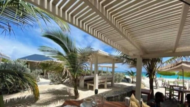 Un área de comedor junto a la playa con una mesa preparada para dos personas, que incluye una bebida, un plato de comida y un vaso de jugo.