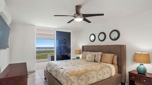 Une chambre avec un grand lit, un ventilateur de plafond, deux lampes de chevet et une vue sur l’extérieur, visible à travers une porte vitrée.