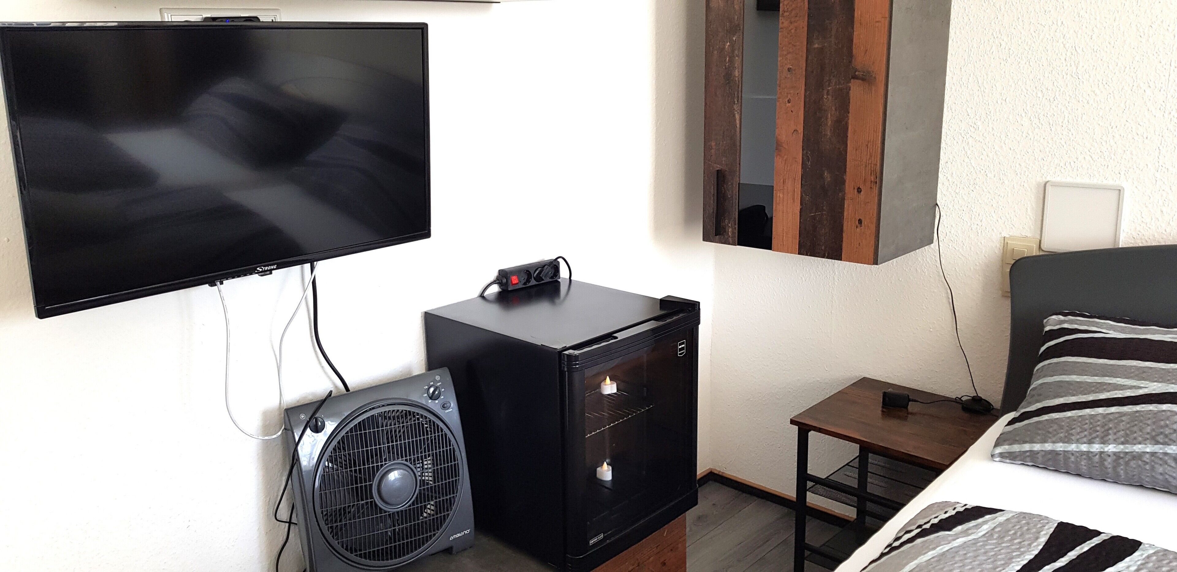 A bedroom with a bed, a TV, a small black cabinet, a fan, and a wooden nightstand.