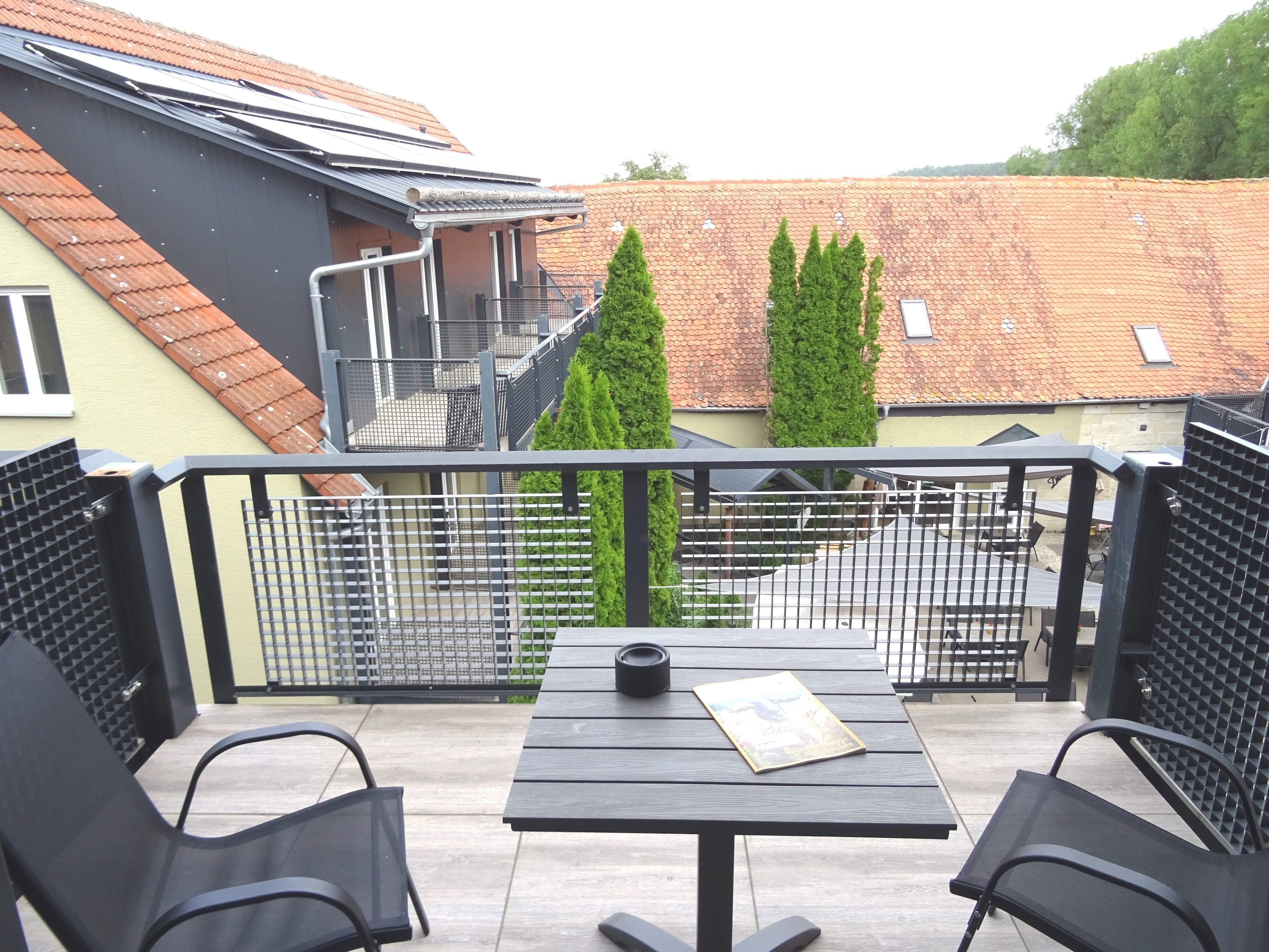 Varanda com mesa e cadeiras, guarda-corrimão de metal e vista para um edifício com telhado de azulejos e duas árvores verdes.