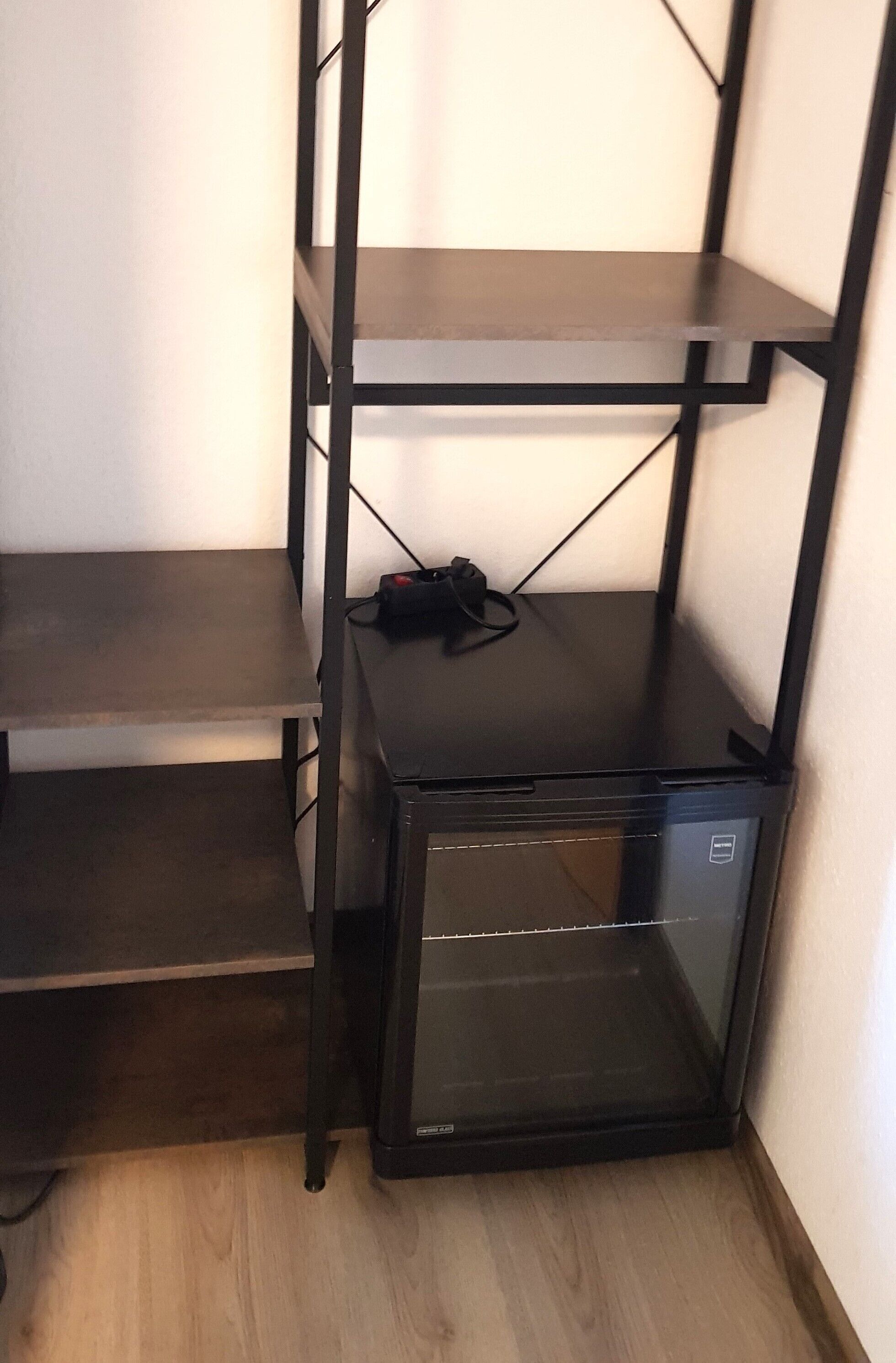A corner with a black metal shelving unit, a small black refrigerator, and a wooden floor.