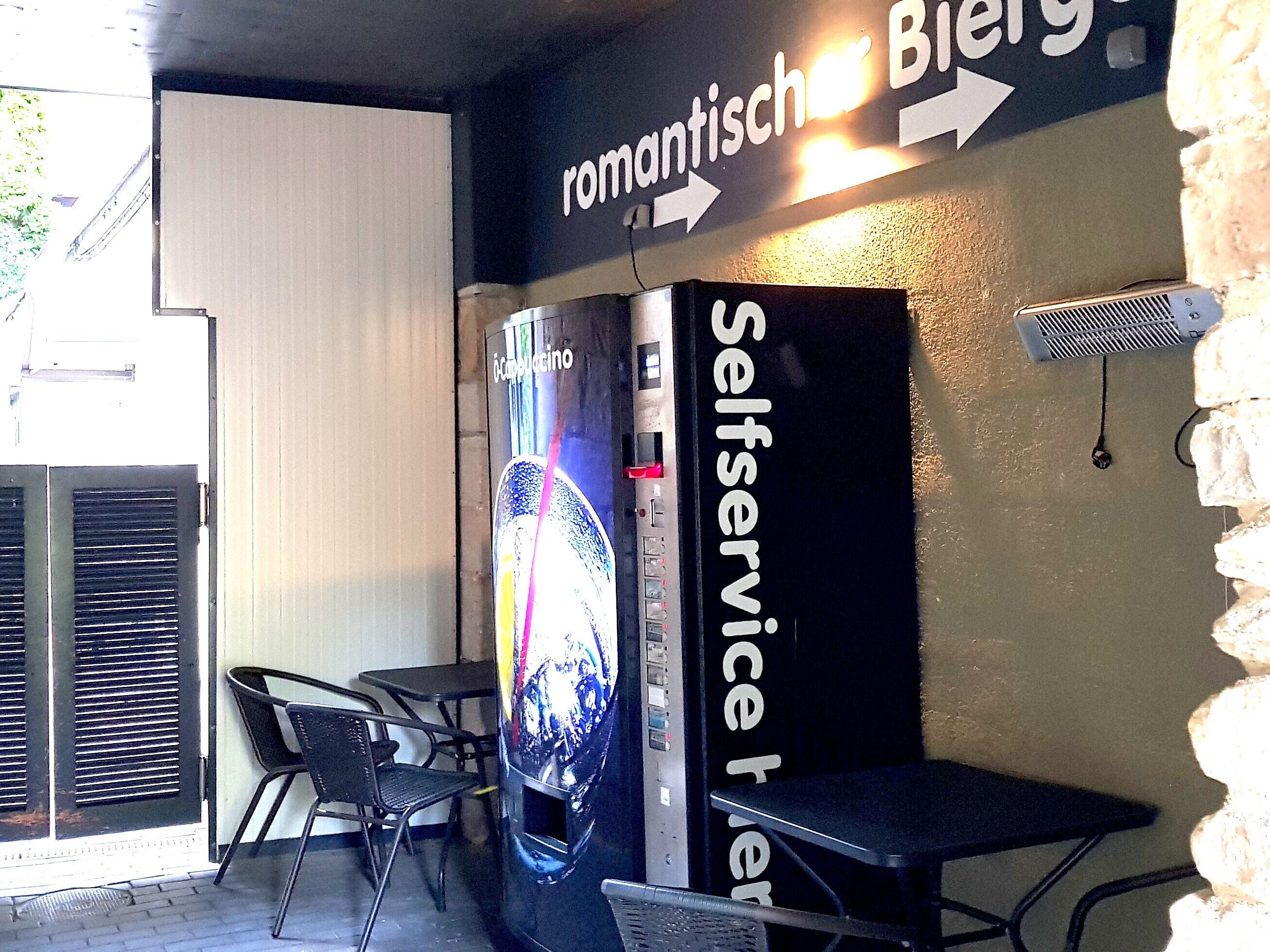 A self-service kiosk with a digital display, a sign indicating 'romantischer Biergarten' with an arrow, and outdoor seating with chairs and tables.