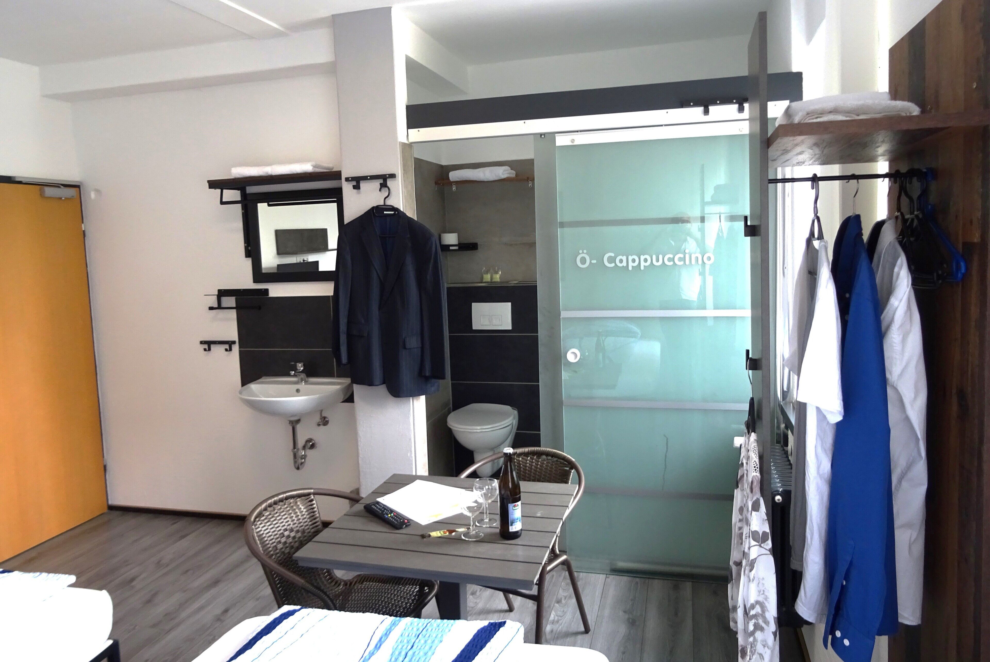 Une chambre d’hôtel avec un lit, une table sur laquelle se trouvent une bouteille, des verres et une télécommande. La salle de bain est équipée d’un lavabo, d’un miroir et d’un porte-serviettes. Des vêtements sont suspendus à un étendoir.