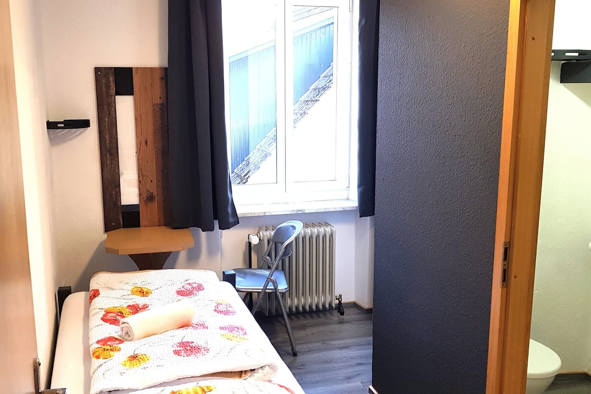 A basic single room with a bed, a chair, a radiator, and a view of a building outside.
