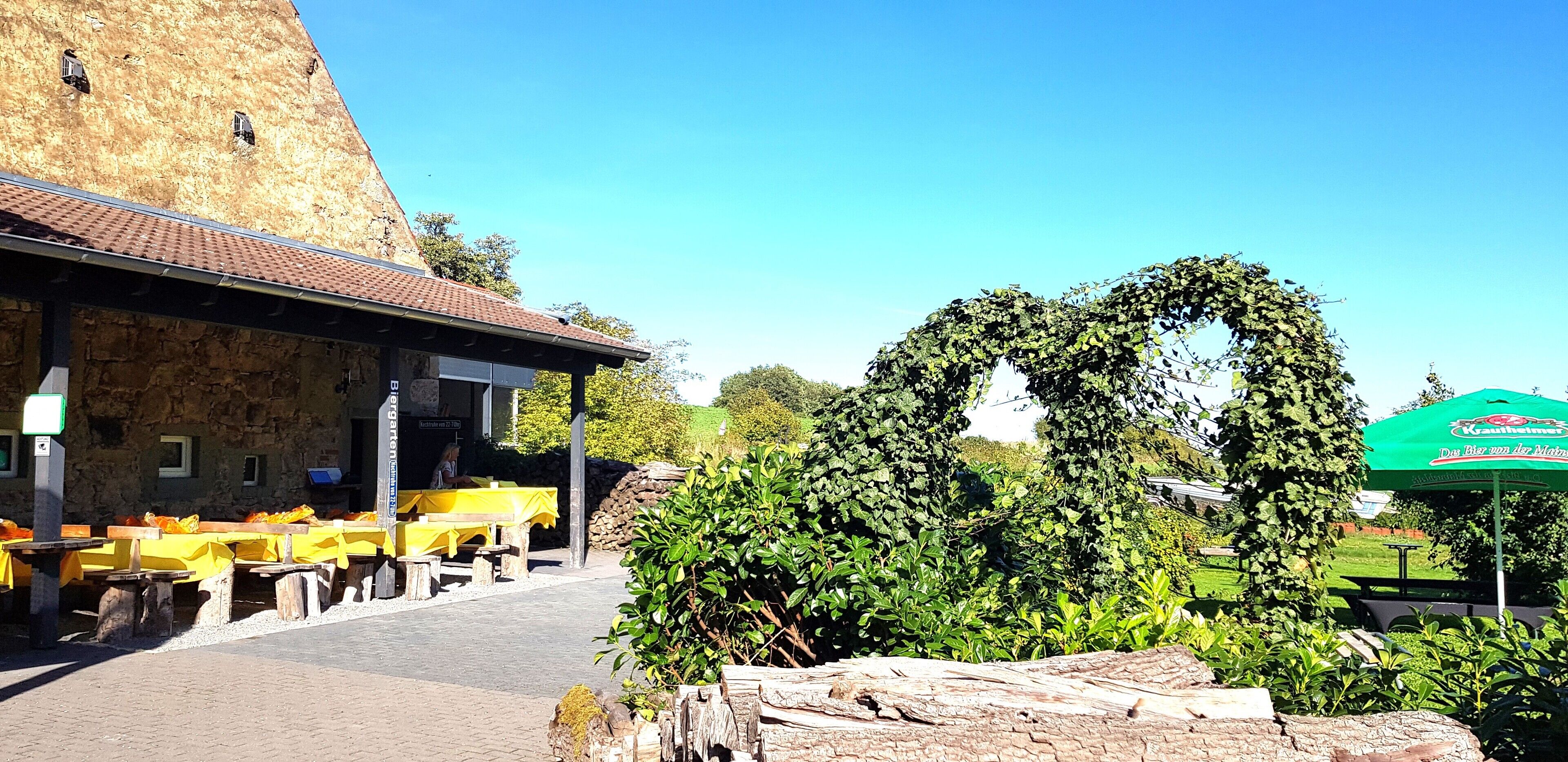 Un espace extérieur aménagé pour les repas, avec des tables et des bancs jaunes, un bâtiment en pierre doté d’un toit en tuiles et un parasol vert.