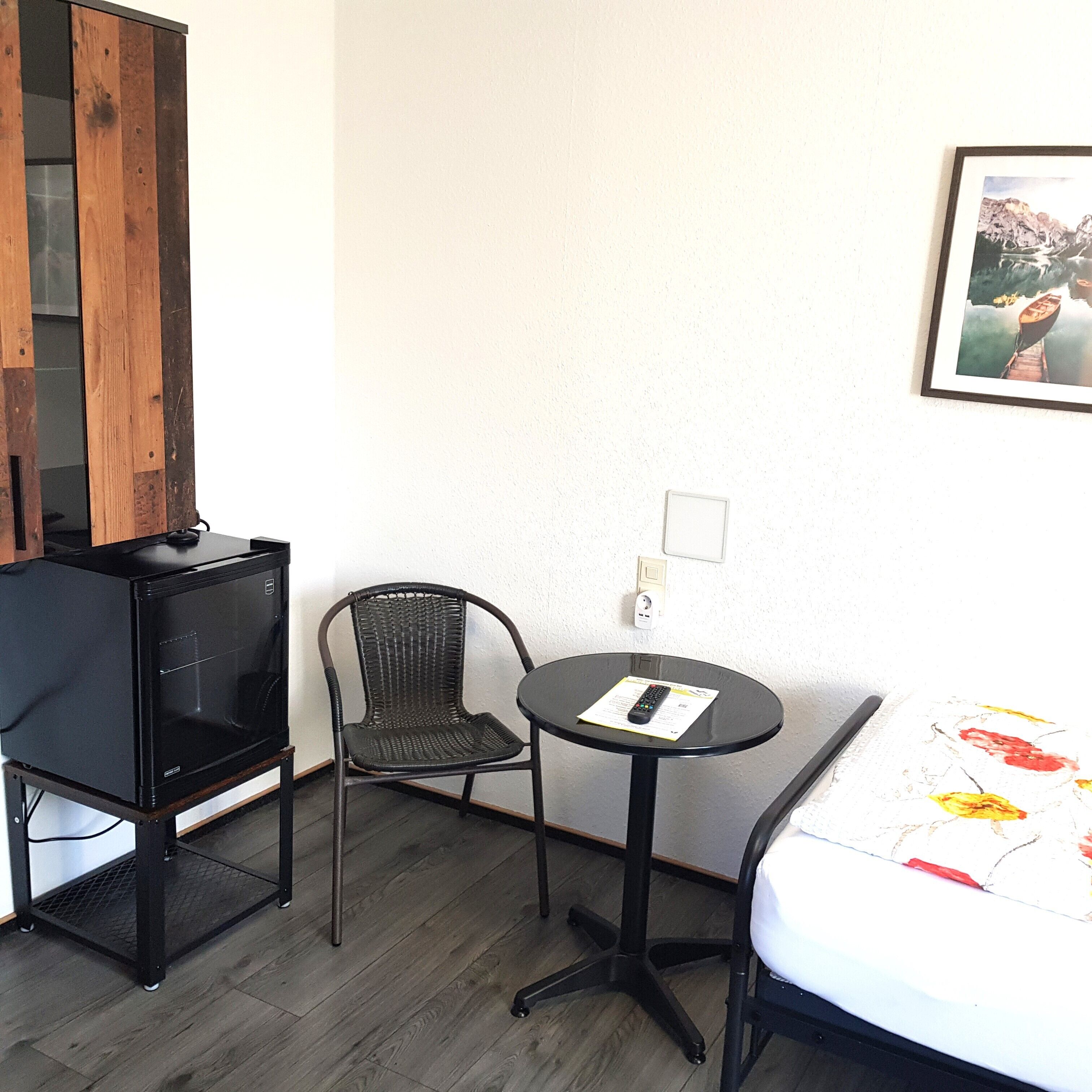 Ein kleines Zimmer mit einem schwarzen Stuhl, einem kleinen runden Tisch mit Fernbedienung und einem Bett mit floralem Überwurf.