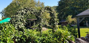 Um jardim com um arco de trepadeira em formato de coração, um gazebo e uma área de recreação.