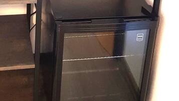A compact black refrigerator in a corner with a wooden floor and a metal shelf.
