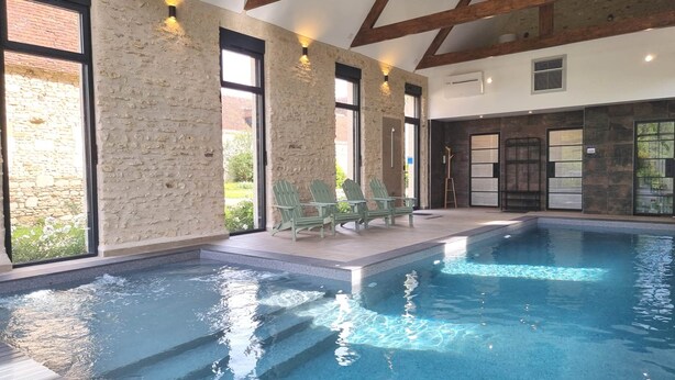 A swimming pool with a stone wall and large windows, surrounded by green chairs.