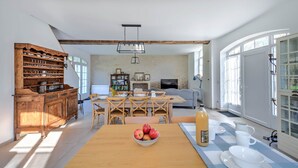 A spacious living room with a wooden dining table, a sofa, and a TV. There is a wooden cabinet with dishes and a fruit bowl on the table.