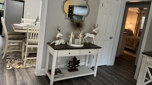 Un área de comedor con una mesa y sillas blancas, un espejo decorativo y un mueble blanco pequeño con cajones.