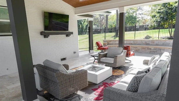 Un espacio al aire libre techado con mobiliario de mimbre, un televisor de pantalla plana y vista a un patio cercado.