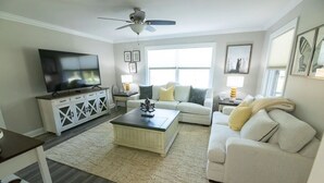 Una sala de estar con televisor, muebles blancos, ventilador de techo y ventanas amplias.