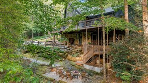 A cabin with a stone staircase leading to a wooden deck, surrounded by trees and a small stream. - 'Make Your Mark' Cozy Sapphire Cabin with Hot Tub (Sapphire)