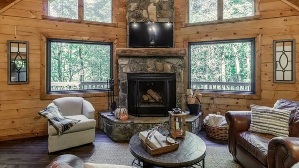 Un acogedor interior de cabaña con chimenea de piedra, paredes de madera, ventanas amplias y una zona de estar cómoda.