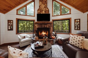 A cozy living room with a fireplace, wooden beams, and large windows. - 'Make Your Mark' Cozy Sapphire Cabin with Hot Tub (Sapphire)