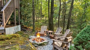 A forested outdoor area with a fire pit, wooden chairs, and a deck. - 'Make Your Mark' Cozy Sapphire Cabin with Hot Tub (Sapphire)