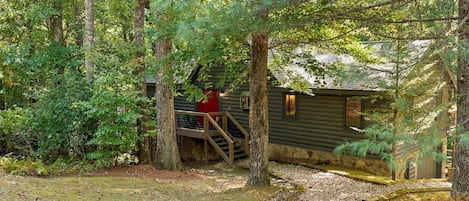 Une cabane avec un escalier en bois menant à une porte rouge, le tout entouré d’une forêt dense.