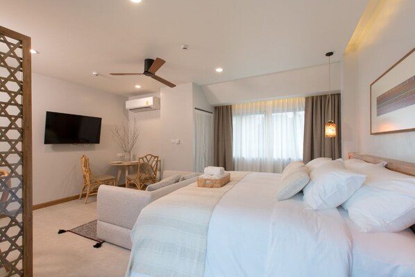 Habitación de hotel con una cama grande, ventilador de techo, televisor de pantalla plana, zona de comedor con mesa y sillas, y ventana con cortinas.