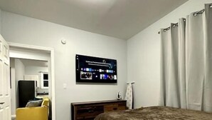 A bedroom with a bed, a TV mounted on the wall, a wooden dresser, and a yellow chair. - Newly 5 Star Rental (New York)