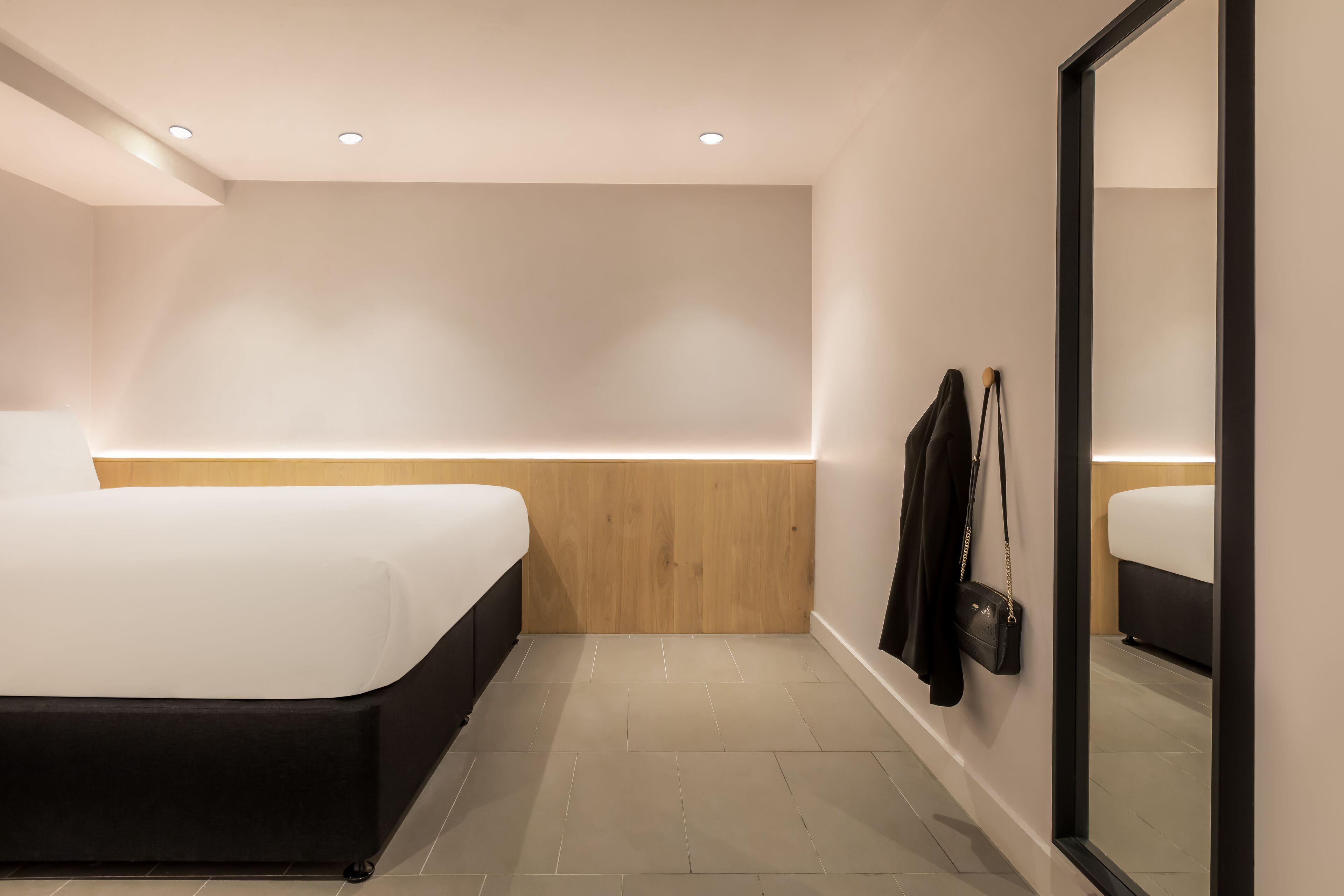 Une chambre d’hôtel moderne avec un grand lit, un mur décoratif en bois, un sac à main noir accroché au mur et un grand miroir.