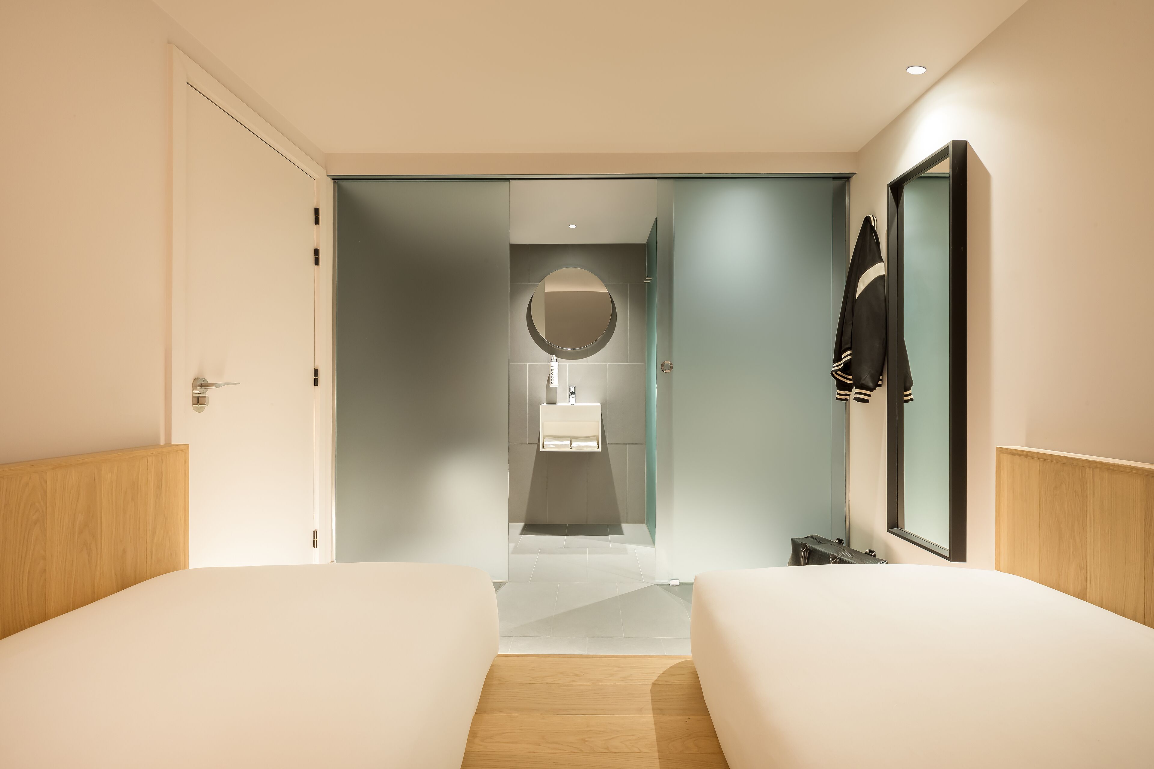 Una habitación de hotel moderna con dos camas, un espejo grande y un baño visible a través de una puerta abierta.