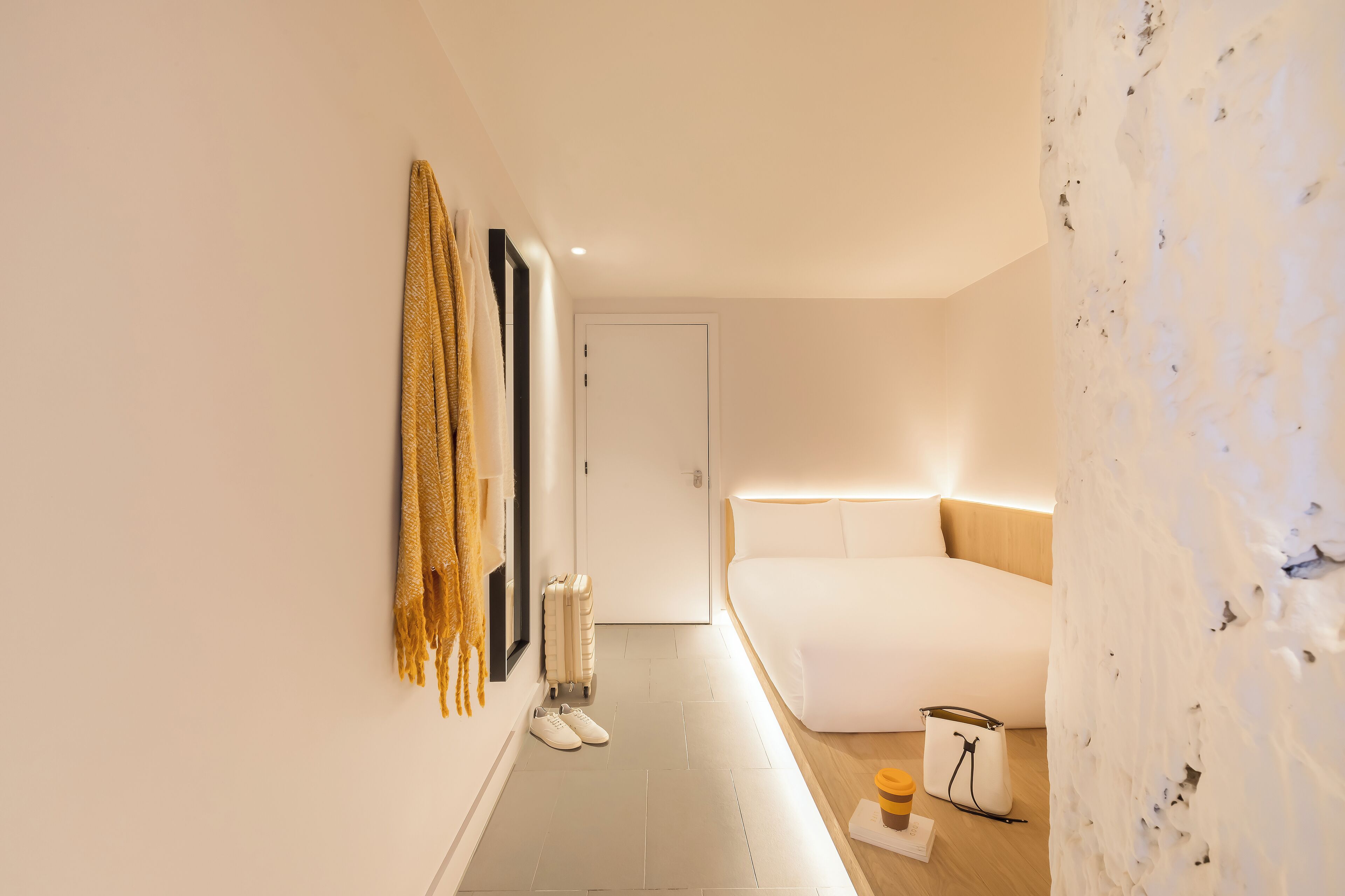 A modern hotel room with a large bed, a white wall with a textured pattern, a door, and a small table with a cup and a bag.