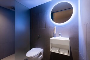 A modern bathroom with a toilet, a sink, and a mirror with a circular light. - Zedwell Underground Hotel Tottenham Court Road (London)