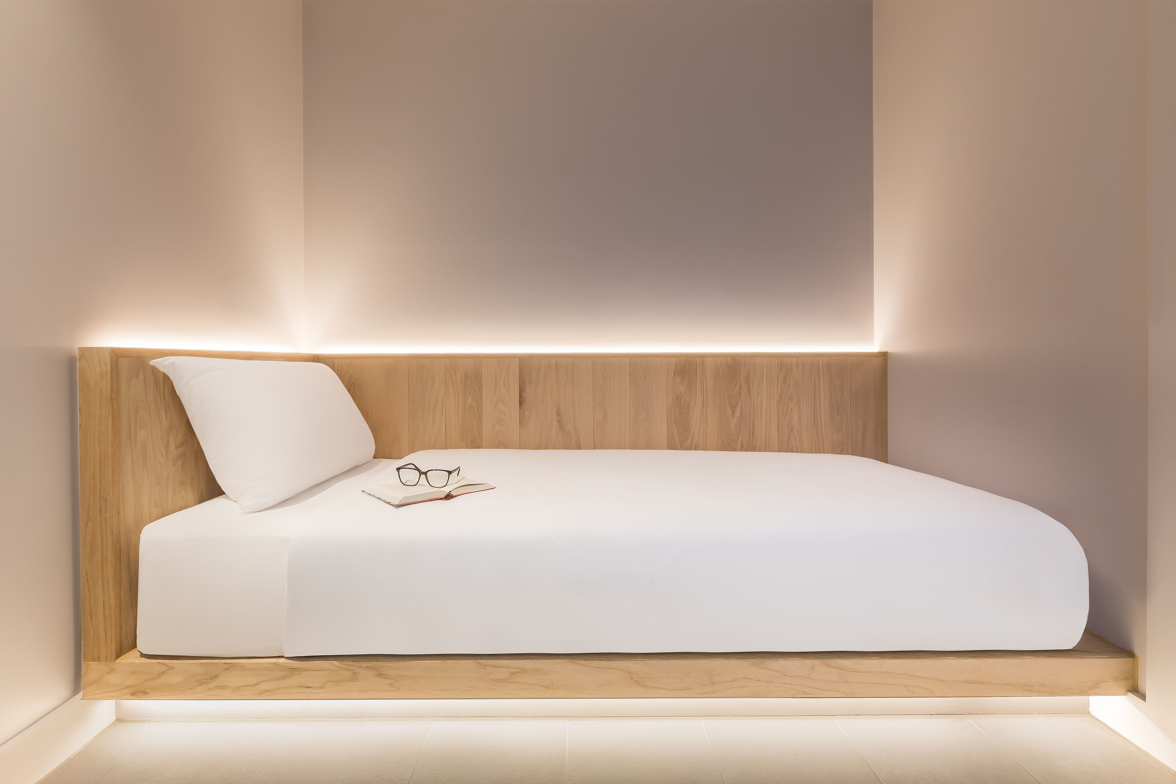 Un dormitorio moderno y minimalista con cabecera de madera, colchón blanco y una mesita de noche con vasos y un libro.