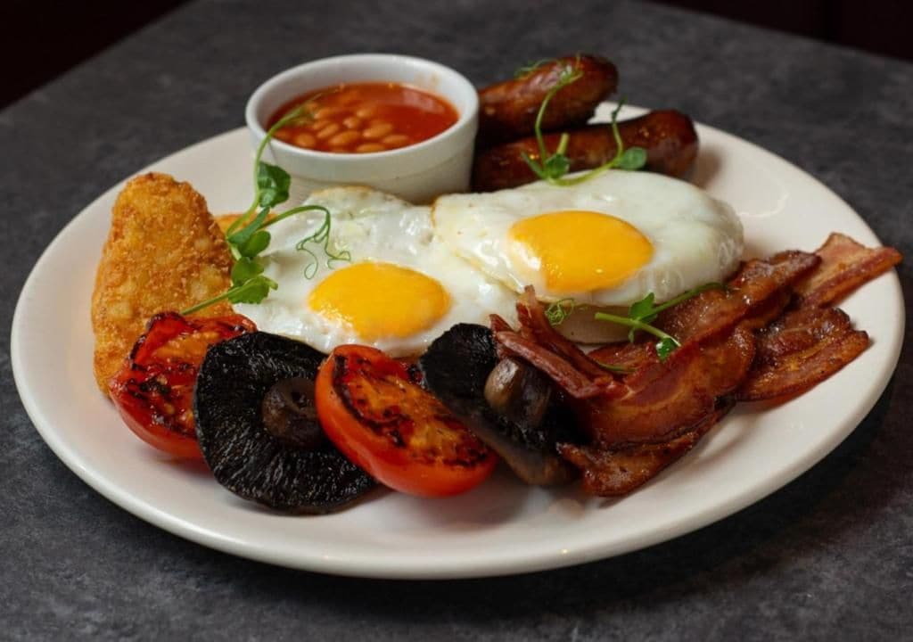 A plate of breakfast food including eggs, bacon, sausages, toast, and grilled vegetables.