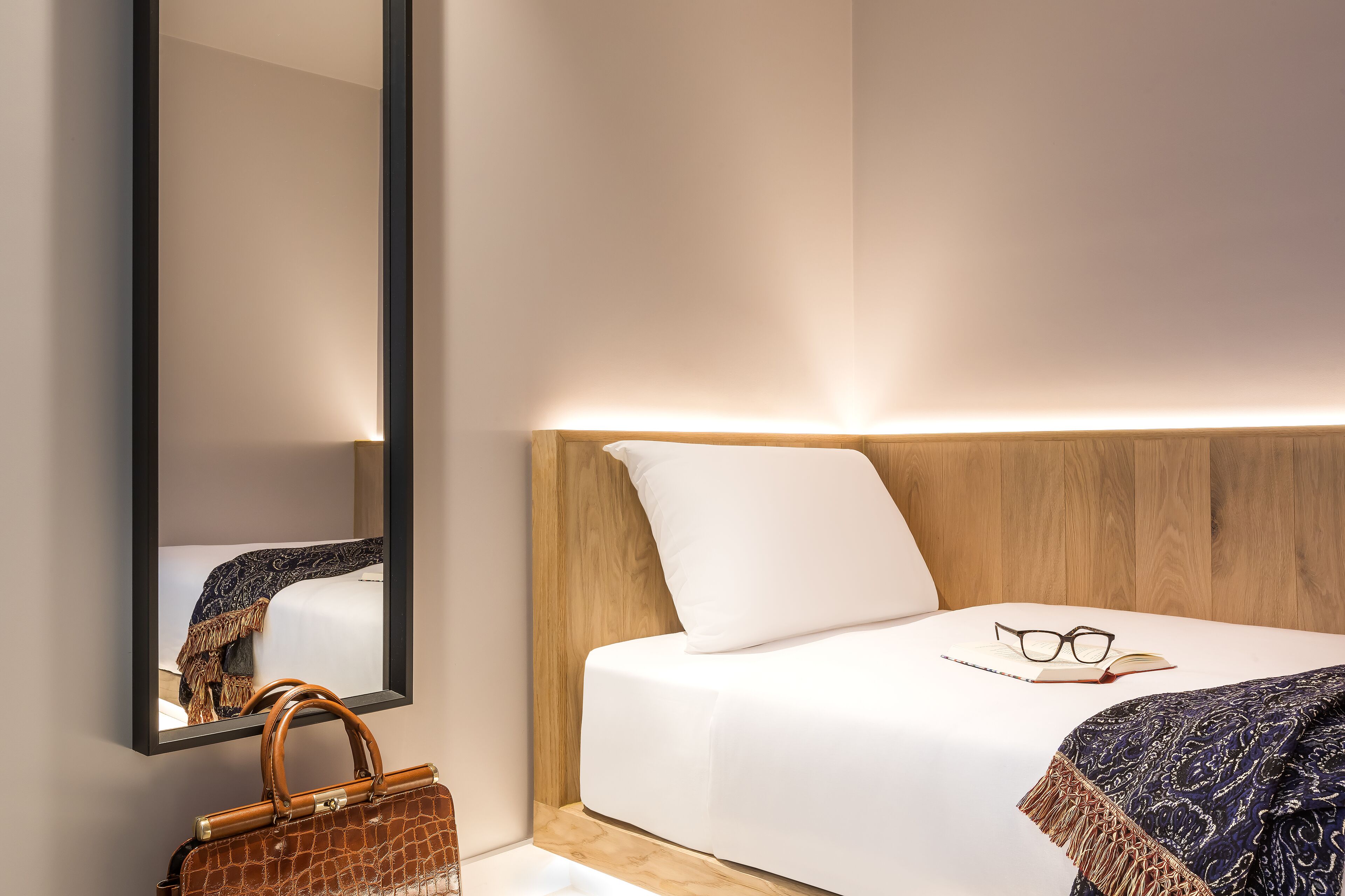 A neatly made bed with a headboard, a bedside table with glasses and a book, and a handbag on the floor.