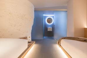 A hotel room with two beds, a round mirror with a light, a sink, and a small table with a bottle and a container.
