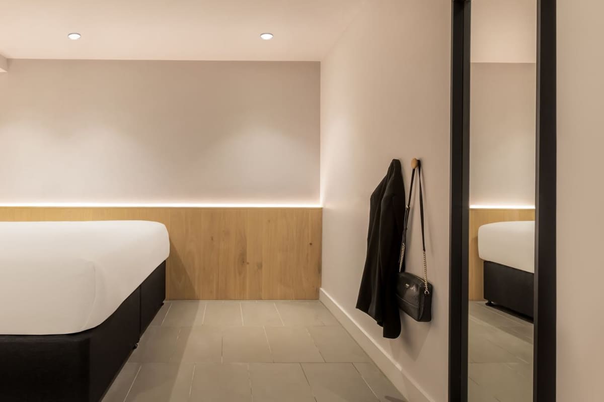 A modern hotel room with a large bed, a wooden accent wall, a black handbag on the wall, and a mirror reflecting the room.