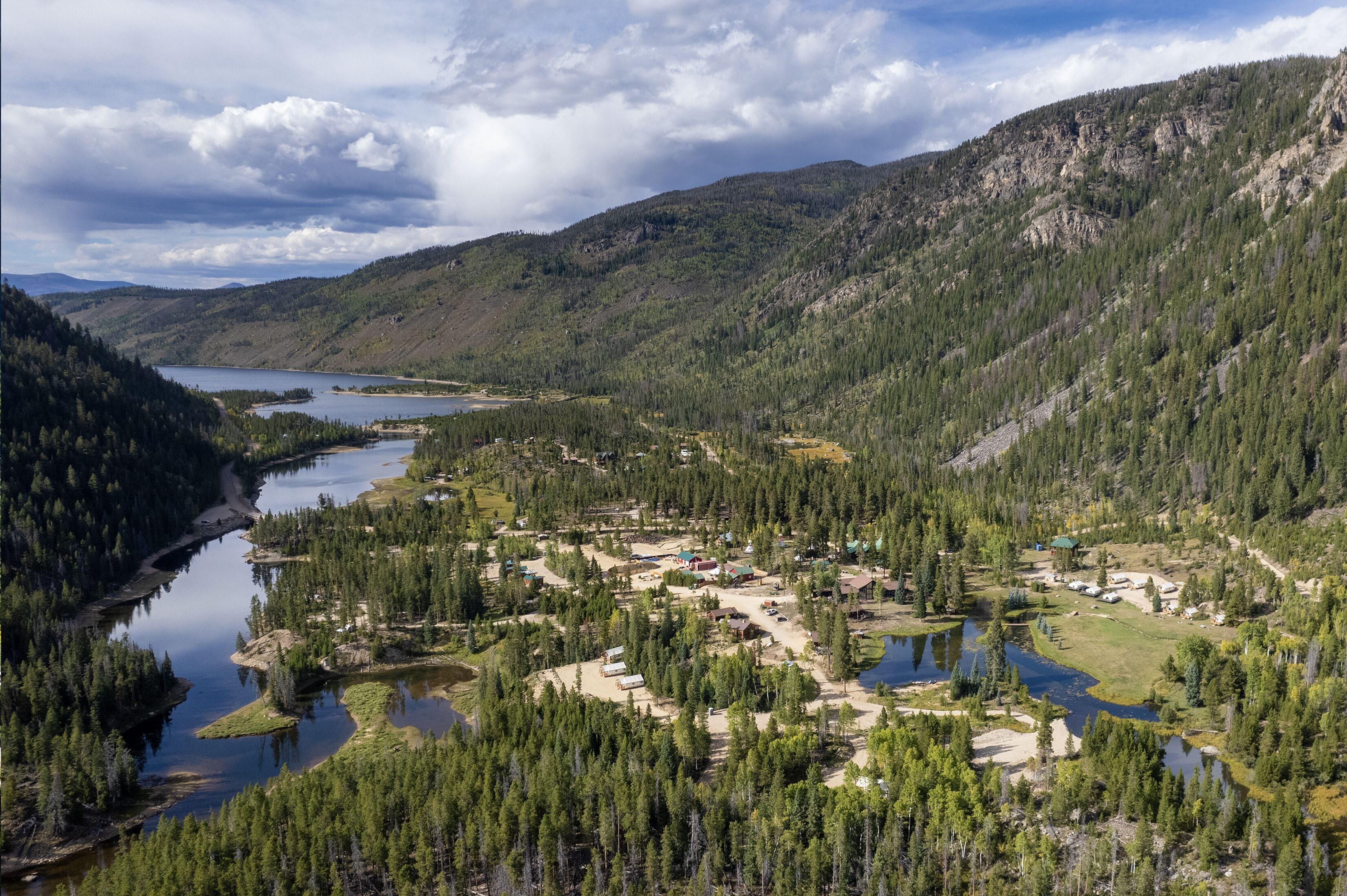 Un paysage montagneux avec une rivière, un terrain de camping et un petit village composé de bâtiments et de tentes.