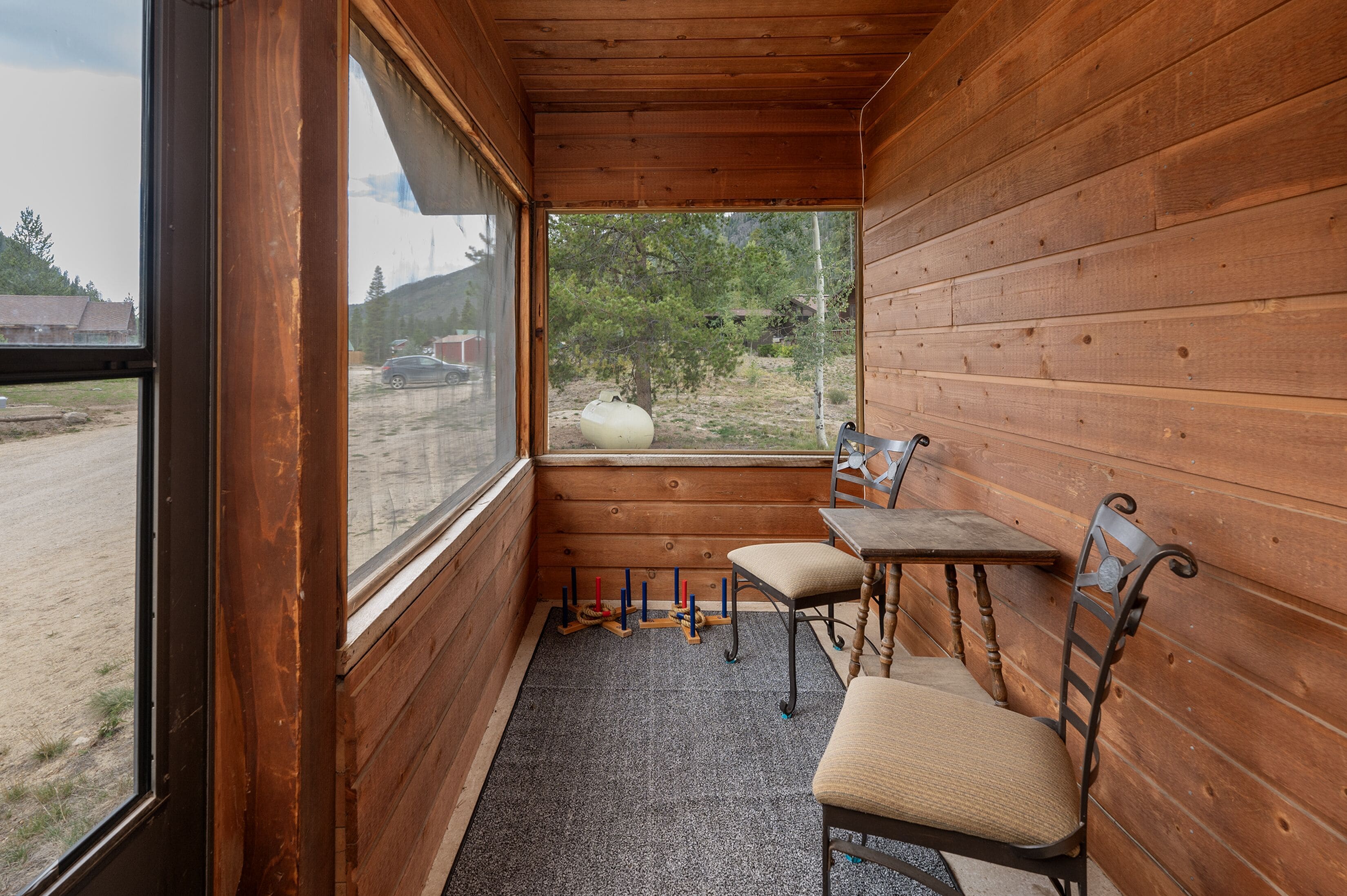 A wooden cabin with a bench and table near a window, a small rug on the floor, and a view of a tree and car outside.