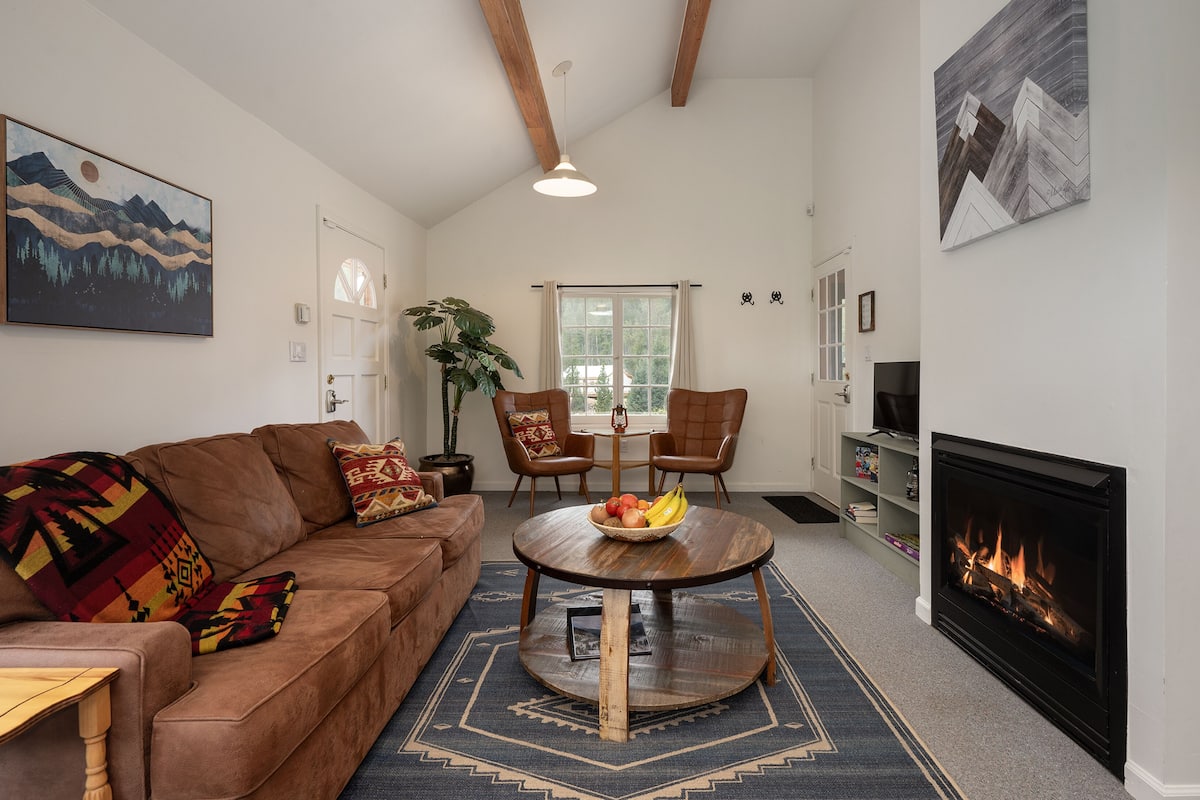 A living room with a brown sofa, a round wooden coffee table with fruits, a fireplace, and a large window.